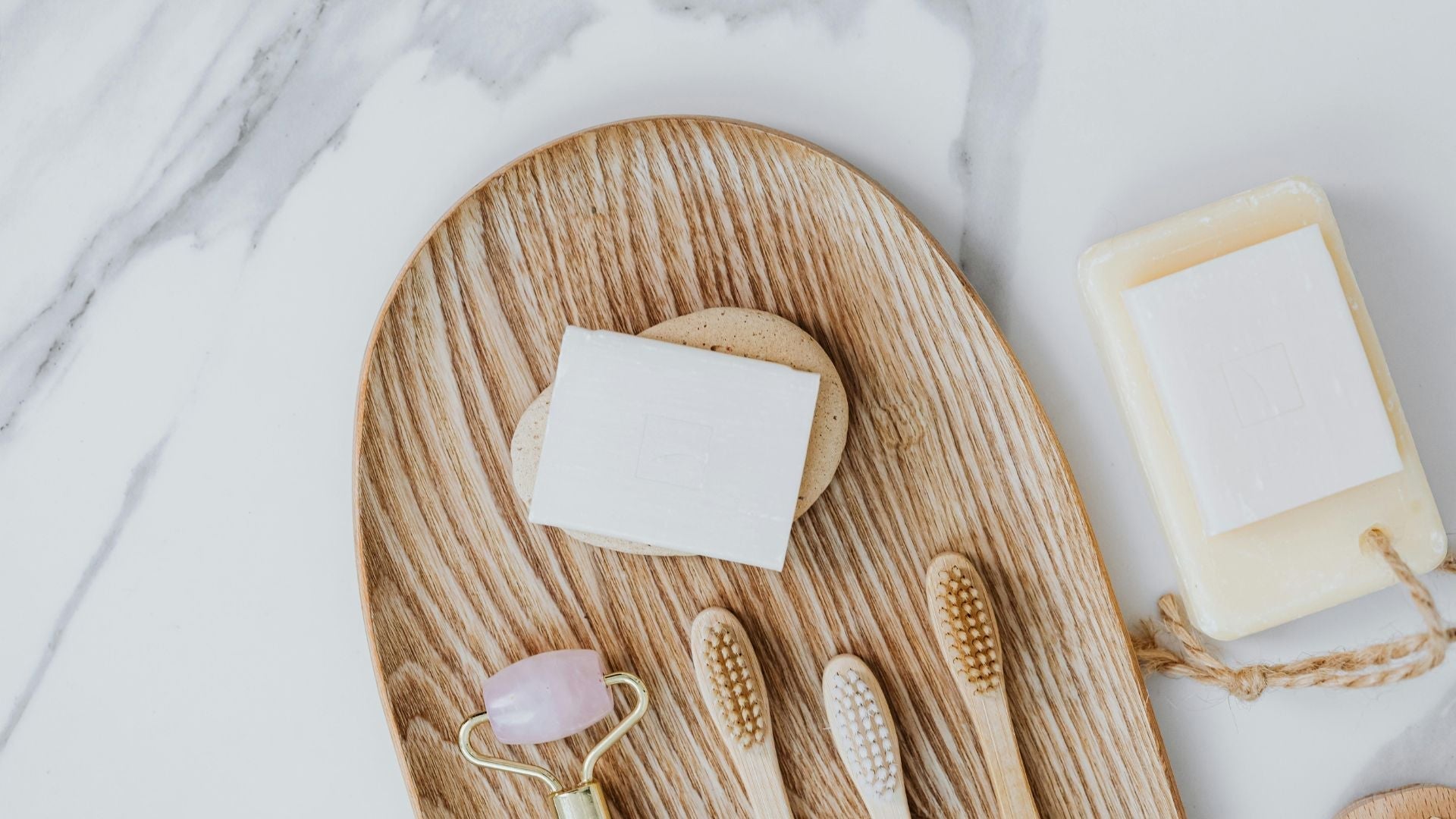 Eco-friendly bathroom essentials on a wooden tray, including bamboo toothbrushes, bar soap, and a rose quartz face roller.