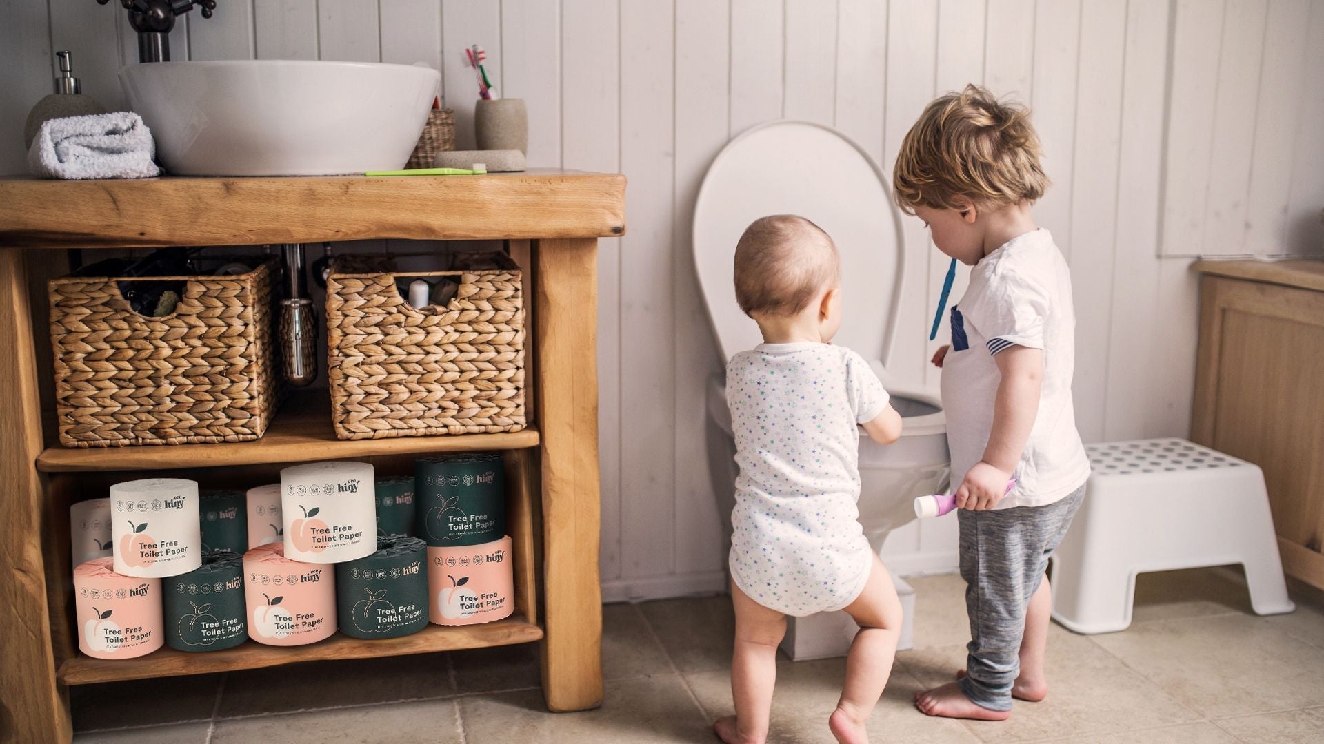 Two young children standing near a toilet in a cozy bathroom with ecoHiny tree-free toilet paper displayed under a wooden sink.