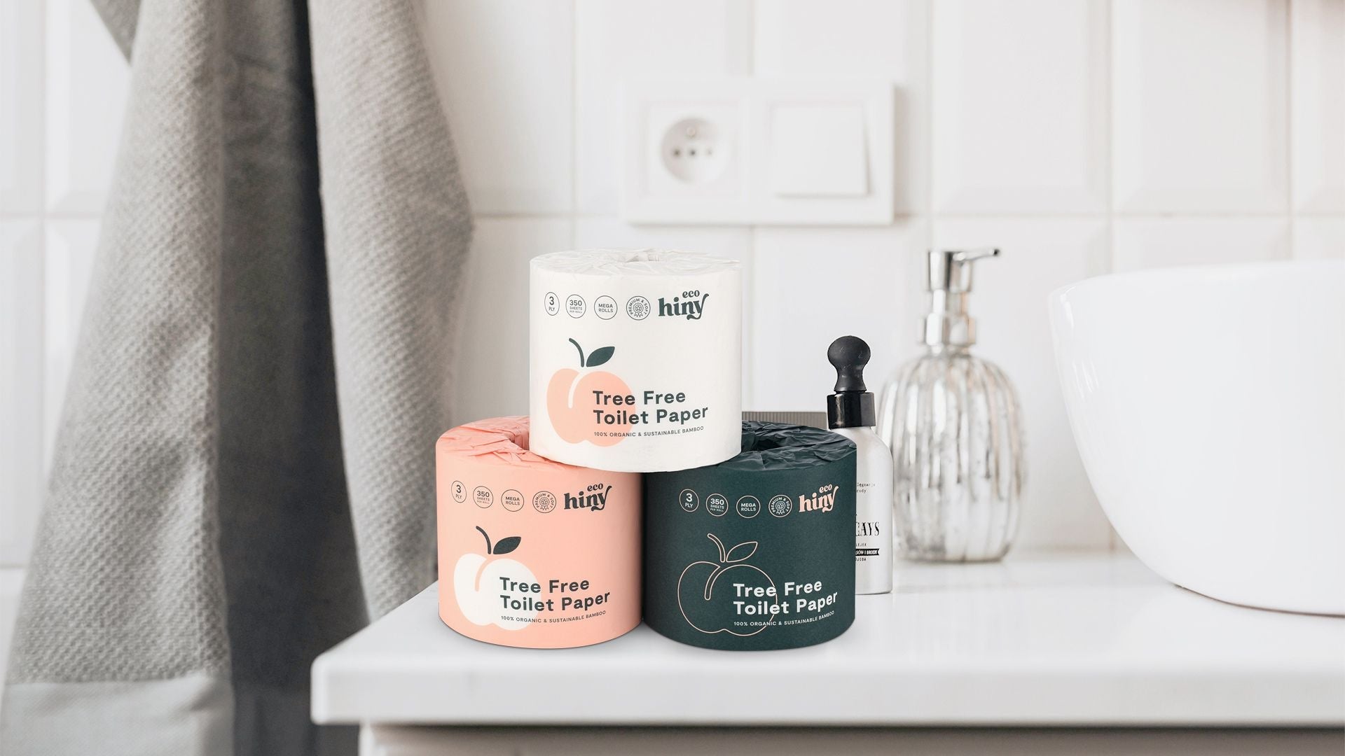 Stack of ecoHiny’s peach, cream, and green bamboo toilet paper rolls in a modern bathroom—showcasing tree-free, plastic-free packaging