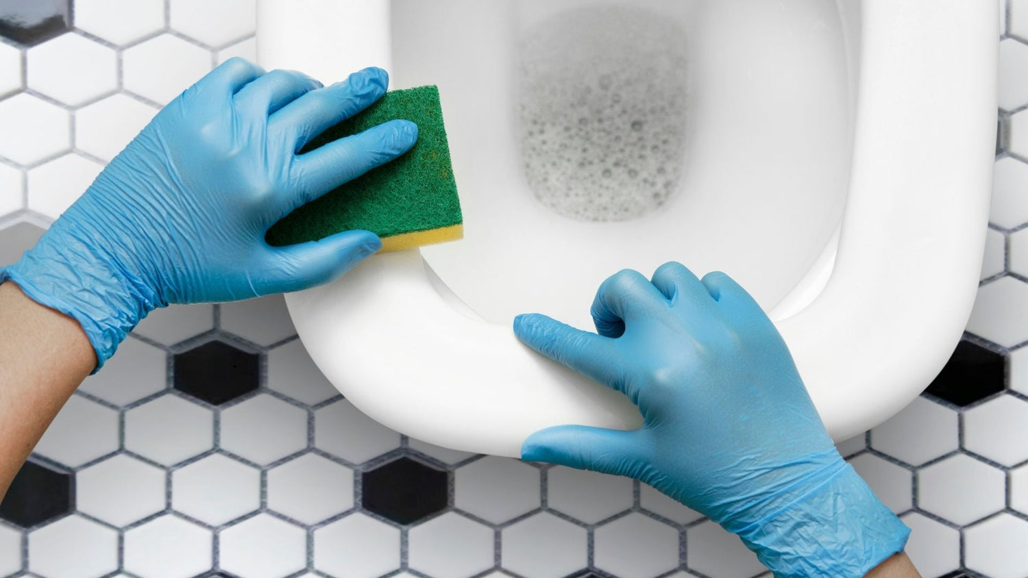 Person wearing blue gloves scrubbing a toilet bowl with a green sponge on hexagon tile floor – eco-friendly bathroom cleaning routine.