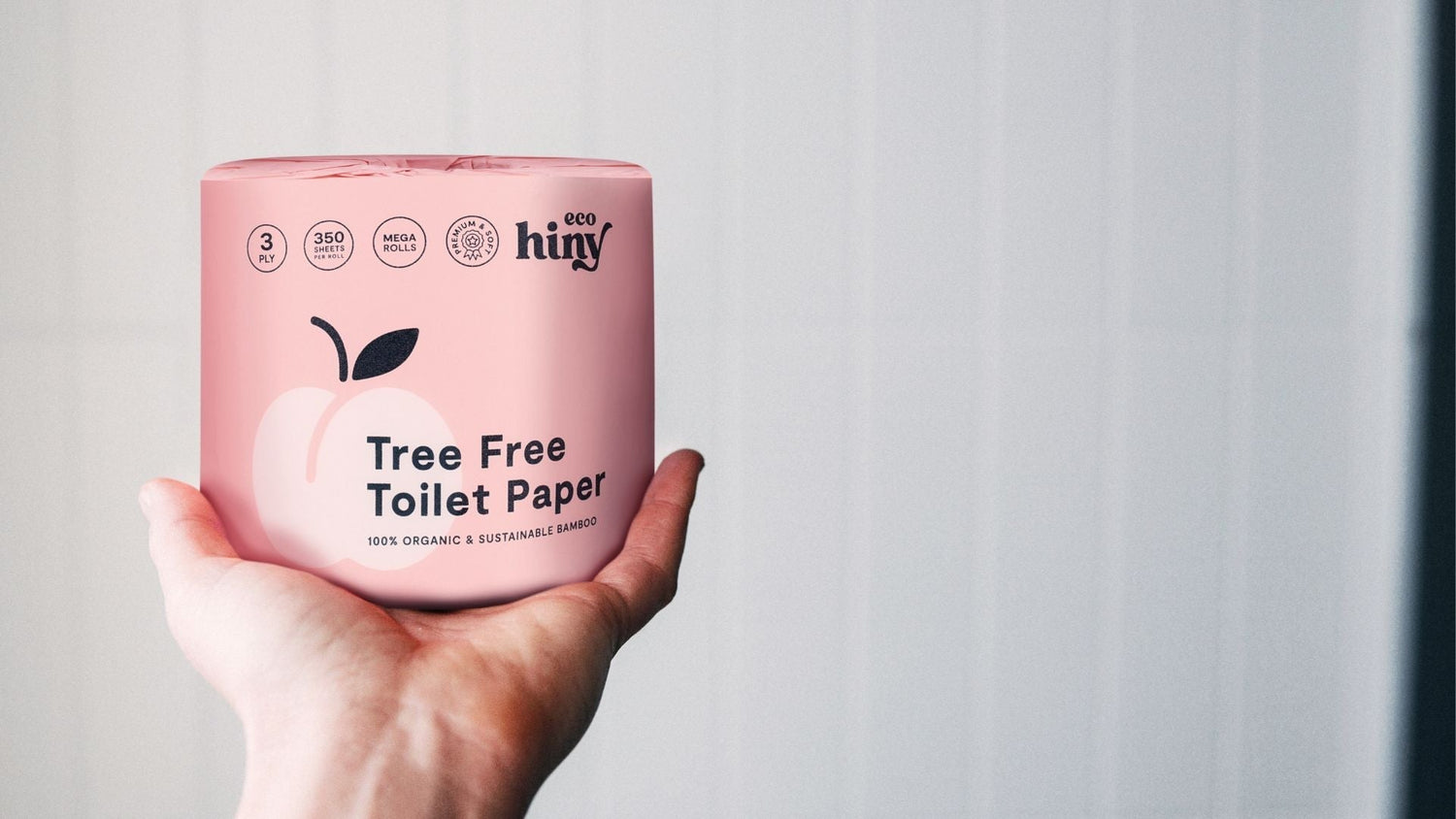  Hand holding a pink-wrapped ecoHiny Tree-Free Toilet Paper made from organic bamboo.