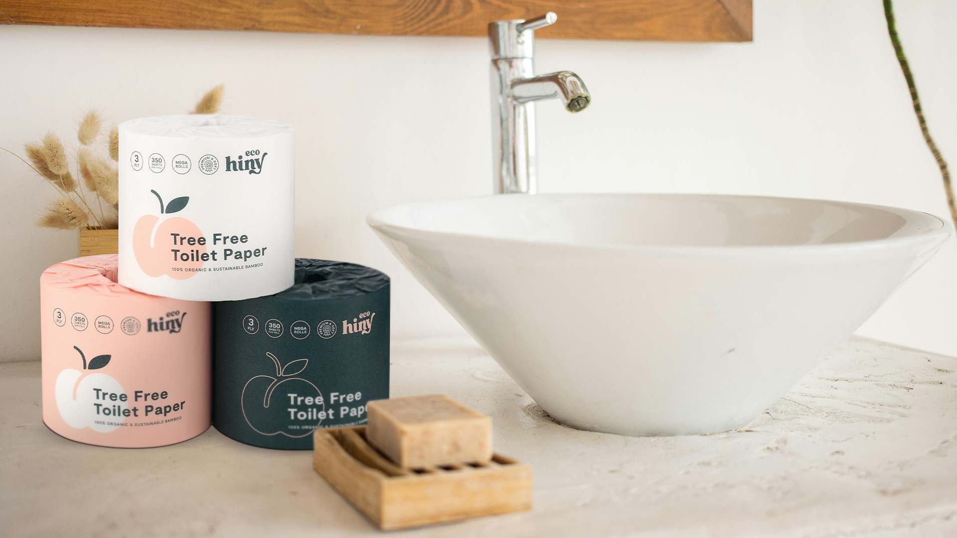 ecoHiny bamboo toilet paper rolls in pink, white, and black packaging placed on a bathroom counter beside a white sink.